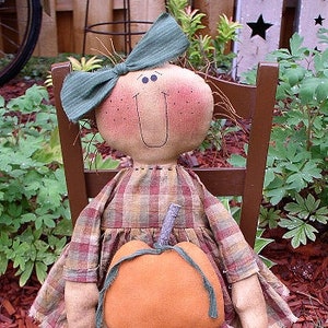 May include: A handmade rag doll wearing a plaid dress and a green bow sits on a wooden chair holding a small orange pumpkin.