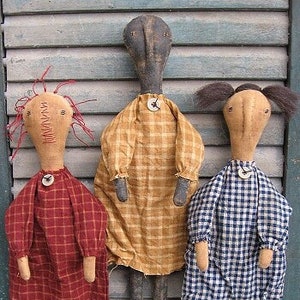 May include: Three handmade rag dolls with different colored plaid dresses. The dolls have painted faces and are standing on a wooden surface. The text "Chestnut Junction" is printed below the dolls.