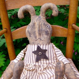 以下が含まれることがあります： 星形のワッペンが付いたストライプのドレスを着た手作りのかやの人形ウサギ。ウサギは、茶色の木製のフレームが付いた籐の椅子に座っています。