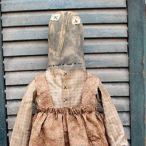 Może przedstawiać: Ręcznie robiona lalka z szmatki w brązowej sukience z białym wzorem pajęczyny. Lalka ma brązową głowę z przyszytym ustami i oczami. Lalka stoi na drewnianym tle.