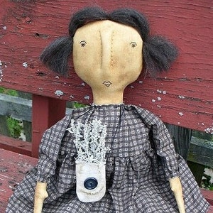 Könnte beinhalten: Eine handgemachte Stoffpuppe mit schwarzen Haaren, die ein braunes und weißes kariertes Kleid trägt und eine weiße Stofftasche mit einem Knopf hält.