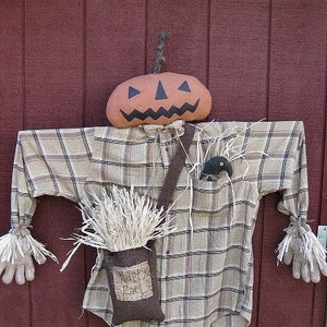 Puede incluir: Un espantapájaros con una camisa a cuadros y una cabeza de calabaza. La calabaza tiene una cara tallada con ojos negros y una amplia sonrisa. El espantapájaros sostiene una bolsa de arpillera que dice "Happy Fall".