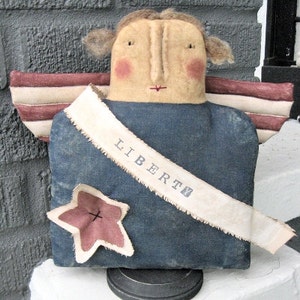 May include: A handmade rag doll angel with red, white, and blue wings. The doll is wearing a blue dress with a white sash that says "LIBERTY". The doll is holding a red star.