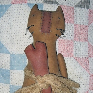 May include: A brown primitive rag doll cat with a red heart in its arms. The cat has a stitched face and is wearing a brown burlap scarf. The doll is sitting on a pink and white quilt.