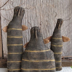 May include: Three handmade primitive style fabric dolls with black and yellow striped bodies and black faces. The dolls are standing on a wooden surface. The dolls are labeled "Chestnut Junction".