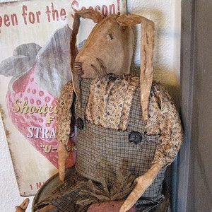 Könnte beinhalten: Eine handgemachte Stoffpuppe in Form eines Hasen, die ein Blumenkleid und eine Latzhose trägt. Der Hase sitzt auf einem Metallbehälter mit dem aufgedruckten Text "Chestnut Junction".