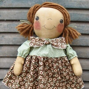 Peut inclure: Une poupée de chiffon faite à la main avec des cheveux bruns en tresses, portant une robe à fleurs verte et marron avec un nœud marron. La poupée est debout sur une surface en bois.