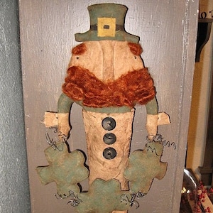 Puede incluir: Un gnomo de fieltro hecho a mano, con un sombrero verde, barba marrón y detalles de trébol. El gnomo sostiene un trébol verde en cada mano. El gnomo está de pie sobre una superficie de madera. El texto "Chestnut Junction" es visible en la imagen.