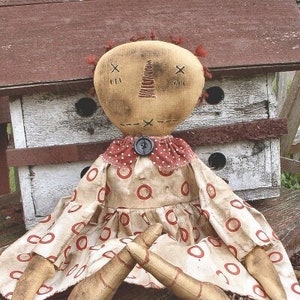 May include: A handmade doll with a tan fabric head and stitched facial features. The doll wears a cream-colored dress with red circle patterns and a red polka-dot collar. The doll's legs are made of wood. The text "Chestnut Junction" is at the bottom.