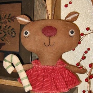Puede incluir: Un reno de peluche marrón hecho a mano que lleva un vestido rojo con lunares y sostiene un bastón de caramelo. La muñeca tiene una cara amigable con ojos negros y una sonrisa cosida. La muñeca está de pie sobre una superficie de madera. El texto "Chestnut Junction" es visible en la parte inferior de la imagen.