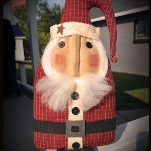 May include: A fabric Santa Claus figure featuring a red and tan plaid hat and body, a white textured brim with a small brown star, black button eyes, a white faux fur beard, and a black belt with a square buckle. The text Chestnut Junction is below.