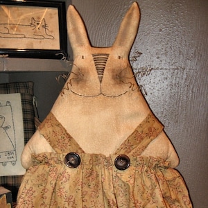 May include: A handmade fabric bunny doll wearing a brown and floral dress with two brown buttons. The doll has a long, pointed nose and a stitched smile. The doll is standing on a wooden surface. The text "Chestnut Junction" is visible at the bottom of the image.