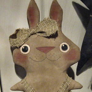 Peut inclure: Une poupée de lapin brun vêtue d'une robe à carreaux et d'un nœud sur la tête. La poupée a des yeux peints et un nez rose. La poupée est assise sur une dentelle blanche. Le texte "Chestnut Junction" est visible en bas de l'image.