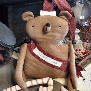May include: A brown plush teddy bear wearing a red and white Santa hat with a white pom-pom. The bear is holding a candy cane and has a red and white ribbon around its neck with the word "Christmas" written on it.