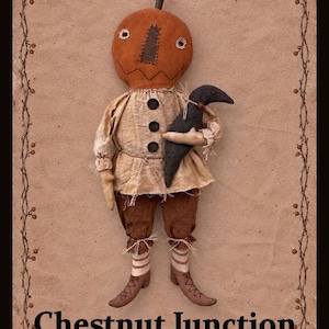 May include: A fabric doll with a round orange pumpkin head, a dark brown stitched nose, and a black crow tucked under one arm. The doll wears a beige shirt with three black buttons, brown trousers, and striped socks with brown shoes. Text reads Chestnut Junction.