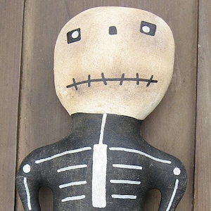 Op de afbeelding: Een handgemaakte stoffen skeletpop met een zwart-wit geschilderd skeletontwerp. De pop houdt een kleine oranje stoffen pompoen vast.