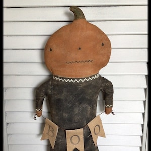 Puede incluir: Una muñeca hecha a mano con forma de calabaza, con un vestido negro y una pancarta que dice "BOO". La muñeca está de pie sobre una superficie de madera blanca.