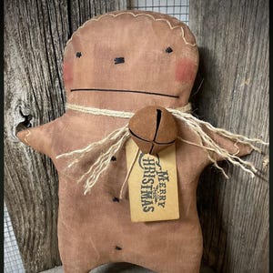 Puede incluir: Muñeco de jengibre de tela marrón hecho a mano con detalles cosidos, mejillas sonrosadas y un collar de cuerda de yute. Tiene una etiqueta de campana que dice "Merry Christmas". La muñeca se exhibe sobre una superficie de madera rústica.