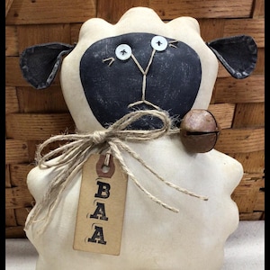 May include: A white sheep doll with a black face and a brown tag that says "BAA". The sheep has a bell attached to its neck and is standing on a woven basket.