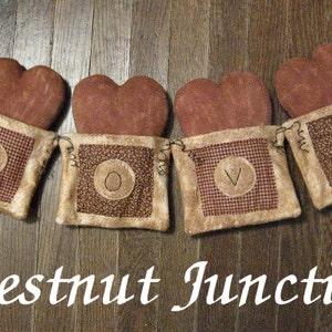 Op de afbeelding: Een rustieke banner met vier bruine vilt harten, elk met een andere letter van het woord "LOVE" geborduurd op een beige vierkant van stof. De banner hangt aan een houten oppervlak.