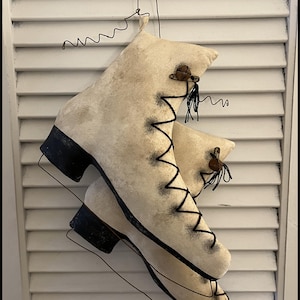Peut inclure: Une paire de patins à glace en tissu blanc avec des garnitures noires et des clochettes brunes suspendues à un fil. Les patins sont vieillis et ont un aspect vintage.