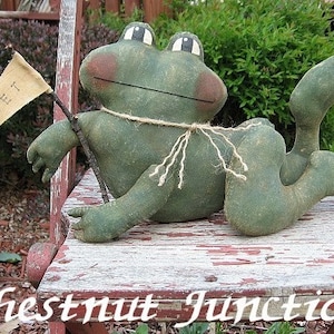 Może przedstawiać: Zielona pluszowa żaba z brązowymi plamkami siedzi na zużytej drewnianej ławce. Żaba trzyma małą flagę z napisem "RIBBET".