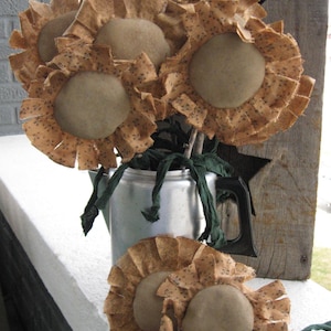 Puede incluir: Un ramo de girasoles de tela marrón con pétalos con volantes y centros marrones. Los girasoles están dispuestos en una jarra plateada con asa negra. La jarra está sobre una superficie blanca.