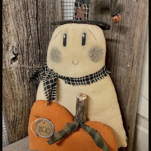 May include: A fabric ghost figurine wearing a black and tan checked hat with a star and a small bell, and a matching checked scarf. The ghost holds an orange fabric pumpkin with a twig stem and a metal tag featuring the text BOO.