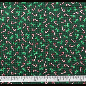 May include: A dark green fabric featuring a repeating pattern of red and white candy canes and green holly leaves with red berries. A white measuring tape is positioned at the bottom, showing a length of approximately 18 inches.