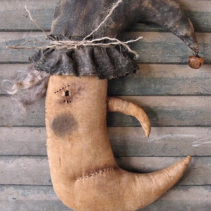 May include: A whimsical handmade doll shaped like a crescent moon with a brown fabric hat and a bell. The doll has a stitched face with a single eye and a brown fabric body with a stitched seam. The doll is displayed against a wooden background.