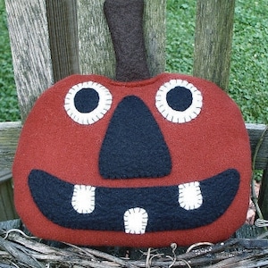 May include: A felt pumpkin with a friendly face. The pumpkin is red with black eyes, a black nose, and a black mouth with white teeth.