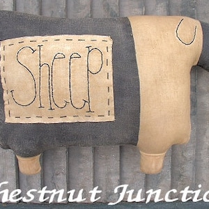 Puede incluir: Un juguete de peluche con forma de oveja marrón y negro con la palabra "Sheep" bordada en un lado. El juguete está sobre una superficie de madera.