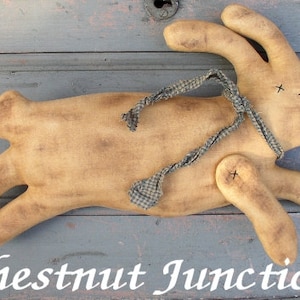 Può includere: Un giocattolo di coniglio primitivo fatto a mano, di colore beige, con un nastro a quadri blu e bianco legato intorno al collo. Il coniglio è sdraiato su una superficie di legno grigia. Il testo "Chestnut Junction" è scritto in bianco sulla superficie.