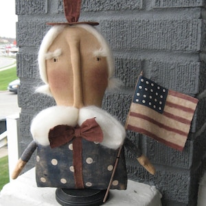 Może przedstawiać: Ręcznie robiona patriotyczna lalka z białą peruką, brązowym kapeluszem i sukienką w niebiesko-białe kropki. Lalka trzyma małą flagę amerykańską.