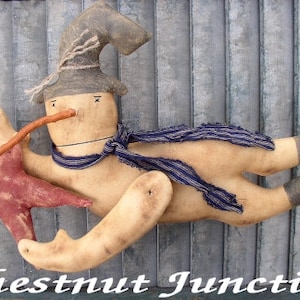 Könnte beinhalten: Eine handgemachte Stoffpuppe eines Schneemanns mit einem braunen Hut, einem blau-weiß gestreiften Schal und einem roten Stern. Der Schneemann fliegt mit einem Stock als Nase durch die Luft.