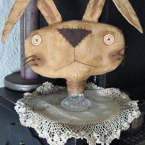 May include: A brown fabric bunny with a black nose and button eyes sits on a white doily on a black surface. The bunny is on a glass pedestal. The text "Chestnut Junction" is visible at the bottom of the image.