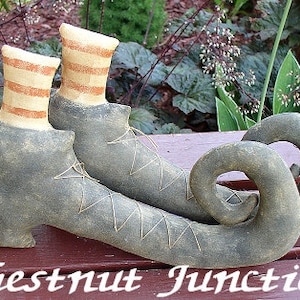 May include: A pair of witch's shoes with curled toes, made of grey fabric with brown stitching. The shoes have red and white striped socks.