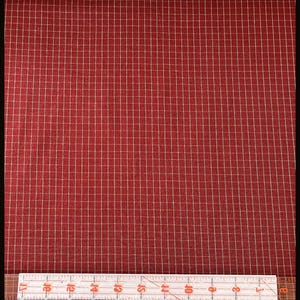 May include: A deep red fabric featuring a fine white grid pattern. A clear plastic ruler is positioned along the bottom edge, showing the grid squares measure approximately 0.25 inches by 0.25 inches across the surface of the textile material.