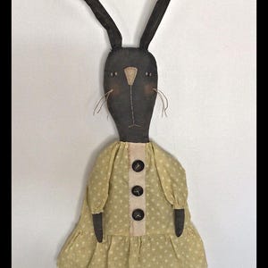 May include: A black rag doll bunny wearing a yellow dress with white polka dots and three black buttons. The bunny has long black ears and a stitched-on face.