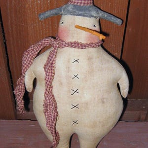 May include: A handmade fabric snowman doll with a red and white checked scarf, a grey hat, and a carrot nose. The snowman has a white body with black stitches for eyes and buttons.