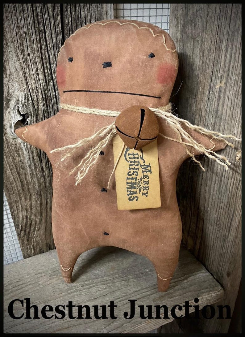 May include: A handmade brown fabric gingerbread man doll with stitched features, rosy cheeks, and a jute twine collar. It has a bell tag that reads "Merry Christmas". The doll is displayed on a rustic wooden surface. The text "Chestnut Junction" is at the bottom.