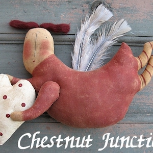 May include: A handmade fabric angel with a red dress, white wings, and a white heart with red polka dots. The angel is holding the heart in its right hand. The angel is flying with its legs outstretched. The text "Chestnut Junction" is written below the angel.