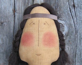 Puede incluir: Una muñeca hecha a mano con cabello castaño, una diadema marrón y un collar de cuentas. La muñeca tiene una cara simple con mejillas pintadas y una pluma en la parte superior de la cabeza.