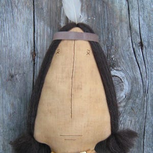 May include: A handmade doll with brown hair, a brown leather headband, and a beaded necklace. The doll is made of fabric and has a simple, stitched face. The doll is displayed against a wooden background.
