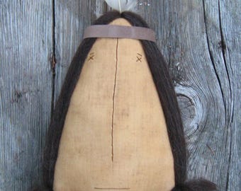 Puede incluir: Una muñeca hecha a mano con cabello castaño, una diadema de cuero marrón y un collar de cuentas. La muñeca está hecha de tela y tiene una cara simple, cosida. La muñeca se muestra sobre un fondo de madera.