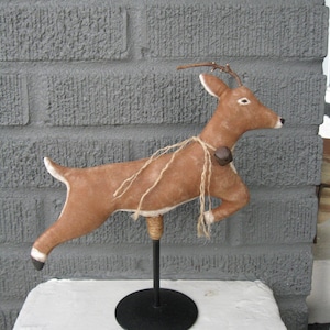 May include: A brown deer figurine with a bell around its neck, mounted on a black metal stand. The deer has a rustic, handmade appearance, with visible stitching and twine accents. The figurine is displayed on a white surface against a grey brick wall.