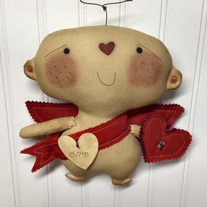 Könnte beinhalten: Handgemachte Valentinstagsdekoration mit einer beigefarbenen Amor-Puppe mit roten Akzenten. Die Puppe hat ein herzförmiges Gesicht, rosige Wangen und ein rotes Herz auf der Stirn. Sie trägt ein rotes Band und hält ein herzförmiges Etikett mit der Aufschrift "CUPID".