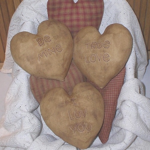 Pode incluir: Almofadas decorativas em forma de coração com mensagens bordadas. Três almofadas castanhas com as inscrições "Be Mine", "True Love" e "I Luv You". Duas almofadas de coração xadrez também são visíveis, tudo sobre uma manta de renda branca.