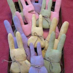 May include: A collection of handmade fabric bunny rabbits in a basket. The bunnies are in various pastel colours, including pink, green, yellow, and purple. Each bunny has a simple, tied bow around its neck.