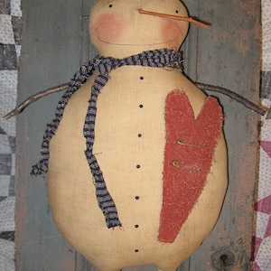 Könnte beinhalten: Ein handgemachter Stoffschneemann mit einem roten Herzen, einem blau-weiß gestreiften Schal und einer Zweignase. Der Schneemann ist aus beigefarbenem Stoff gefertigt und hat schwarze Knopfaugen.
